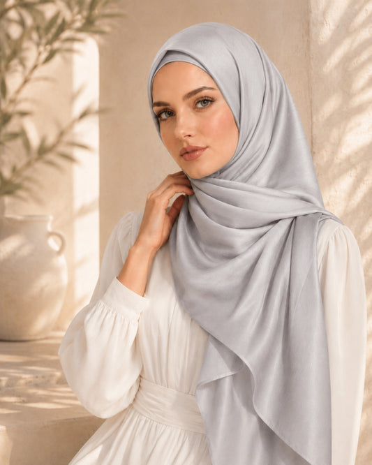 Woman wearing a light gray hijab and white dress against a neutral background.