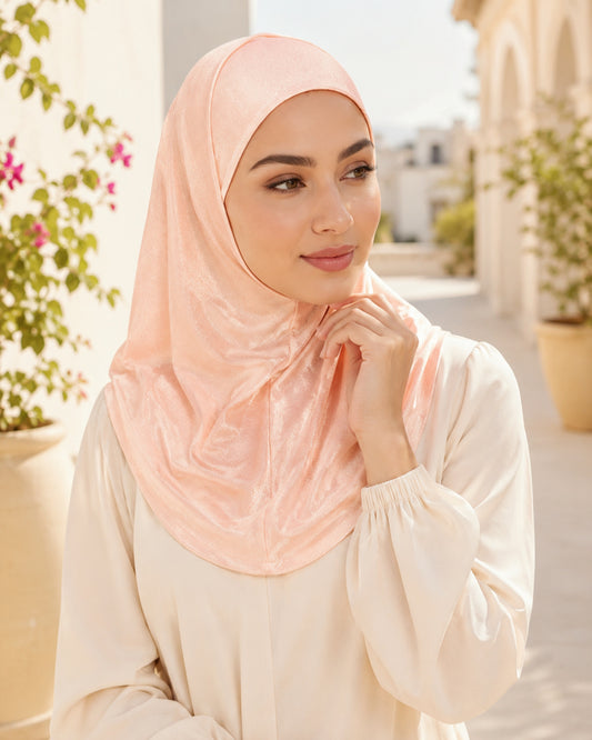Woman wearing a light pink hijab and beige top outdoors with plants in the background