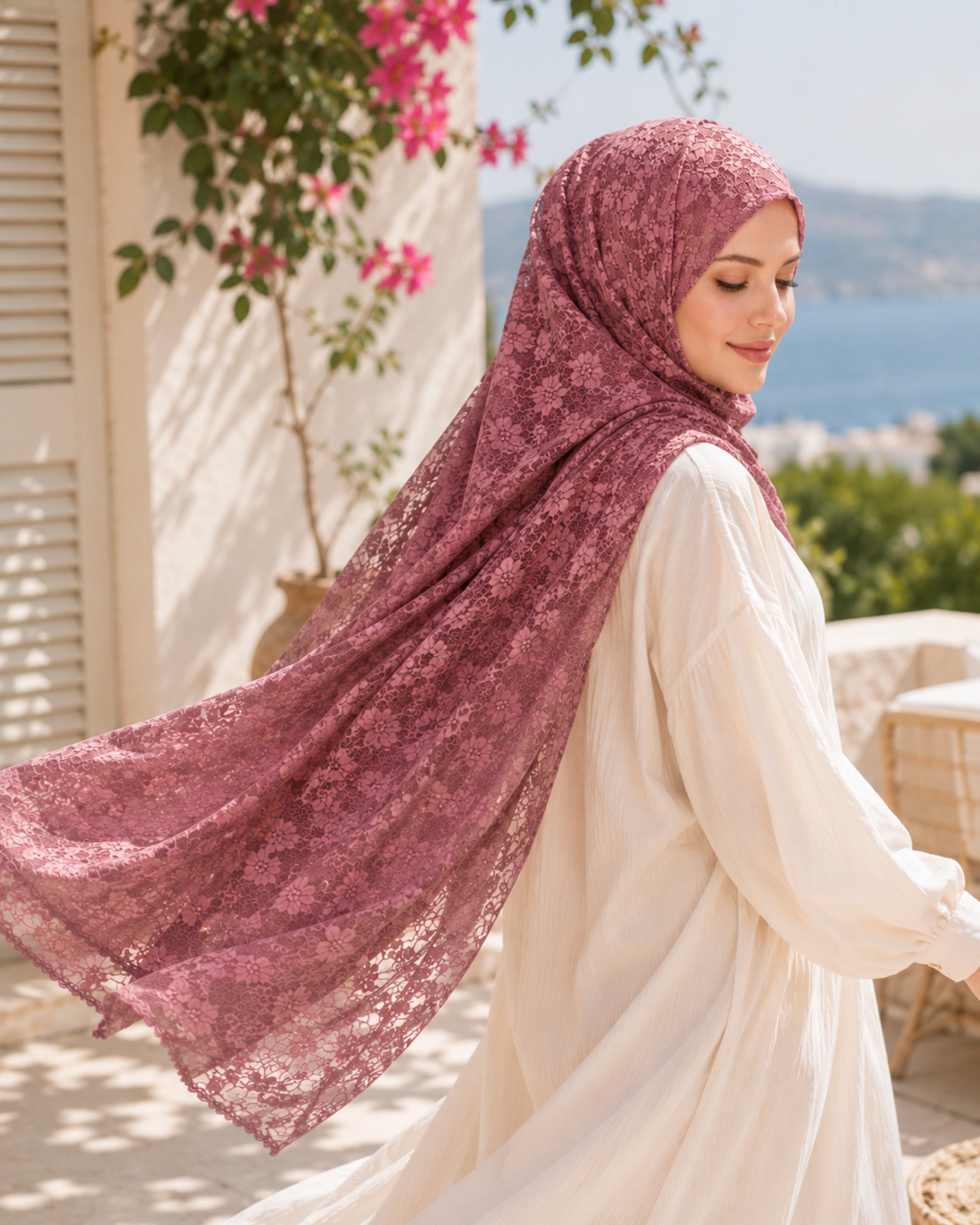 Woman wearing a Mulberry Lace Shawl hijab with floral lace detailing, flowing in the breeze on a sunlit summer terrace.