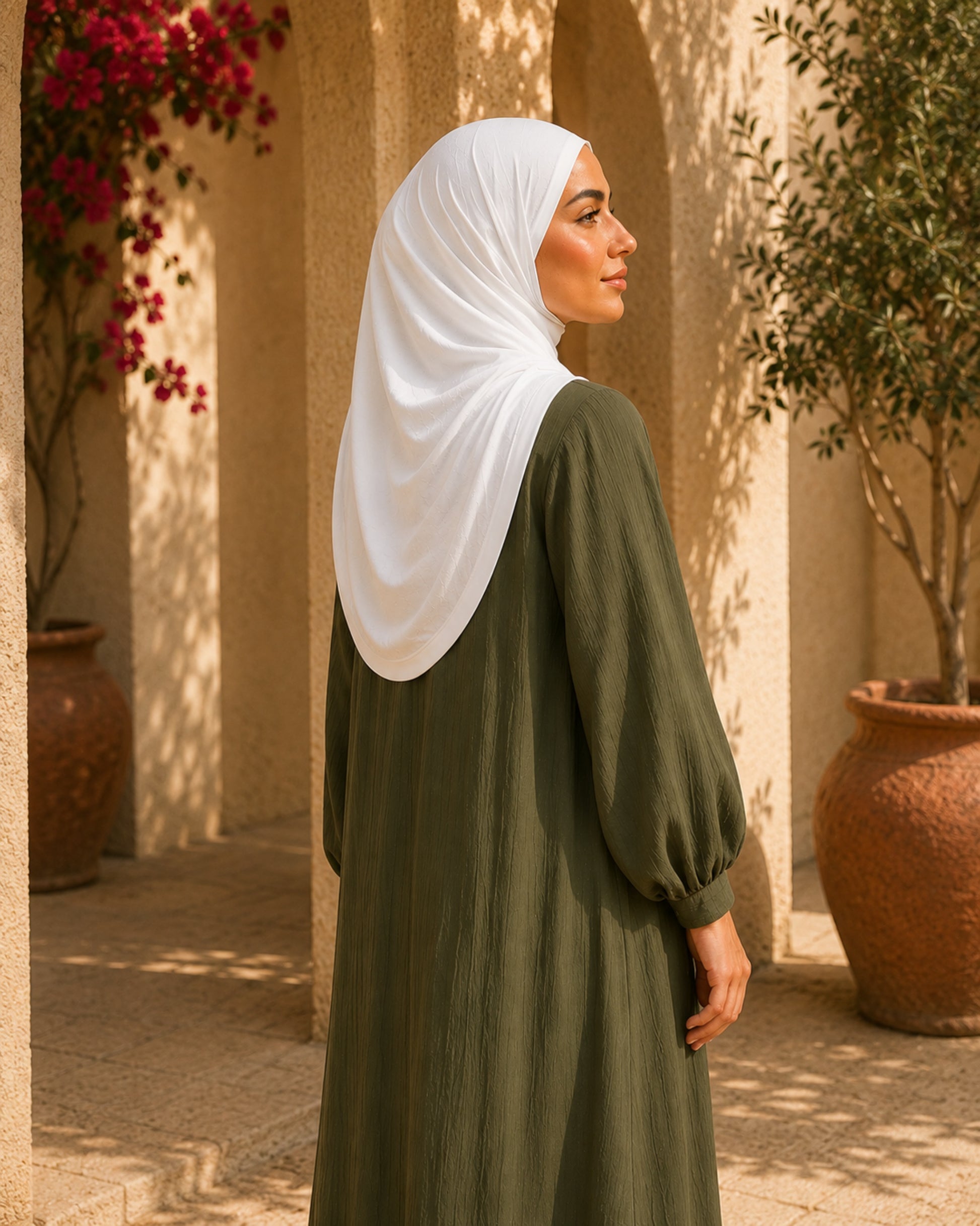 Woman wearing a white hijab and green dress standing in a sunlit outdoor setting with plants and pots.

