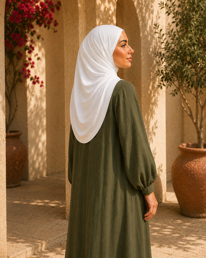 Woman wearing a white hijab and green dress standing in a sunlit outdoor setting with plants and pots.

