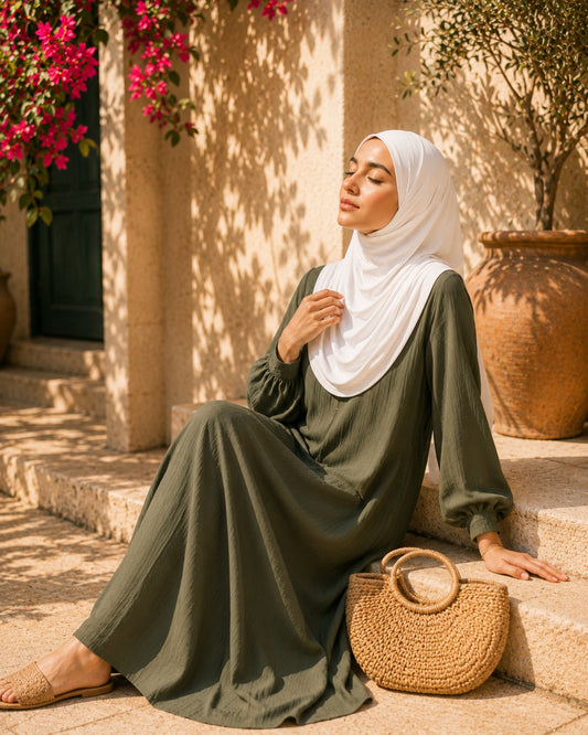 Woman in a green dress and white hijab sitting on steps with a woven bag, surrounded by flowers and a tree.

