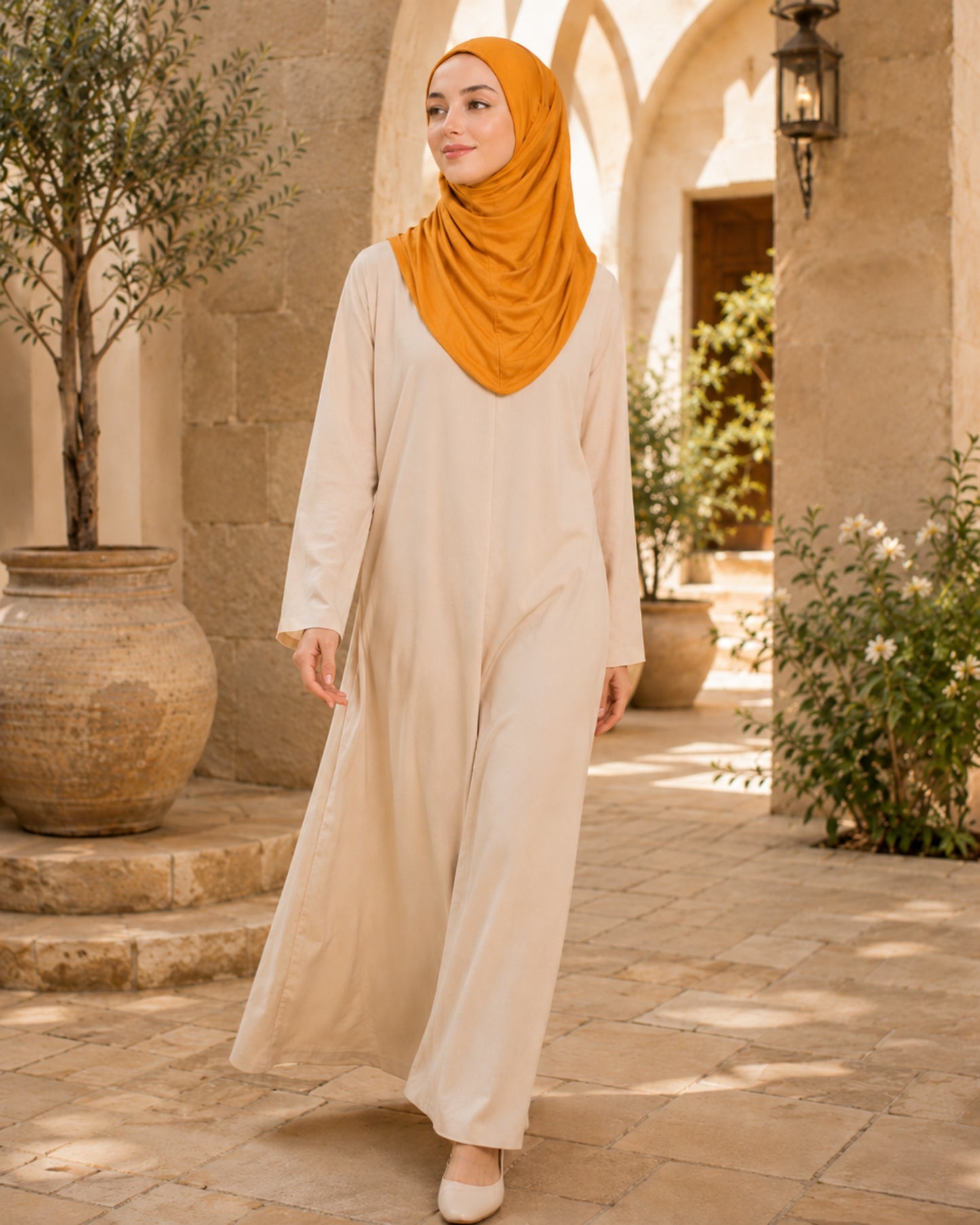Woman wearing a beige outfit and orange hijab standing in an outdoor setting with plants and stone architecture.

