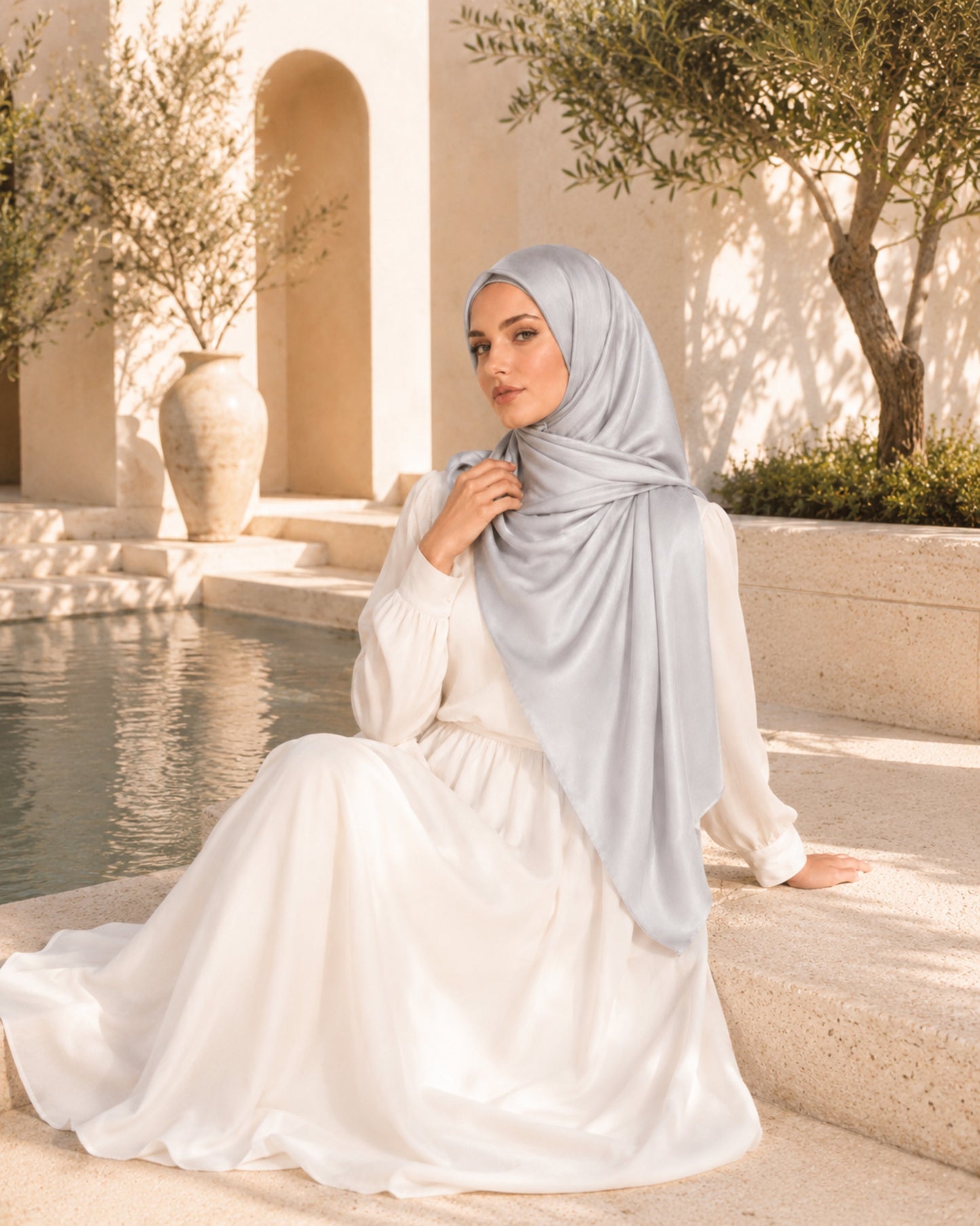Woman in a white dress and light blue hijab sitting by a pool with a scenic background.