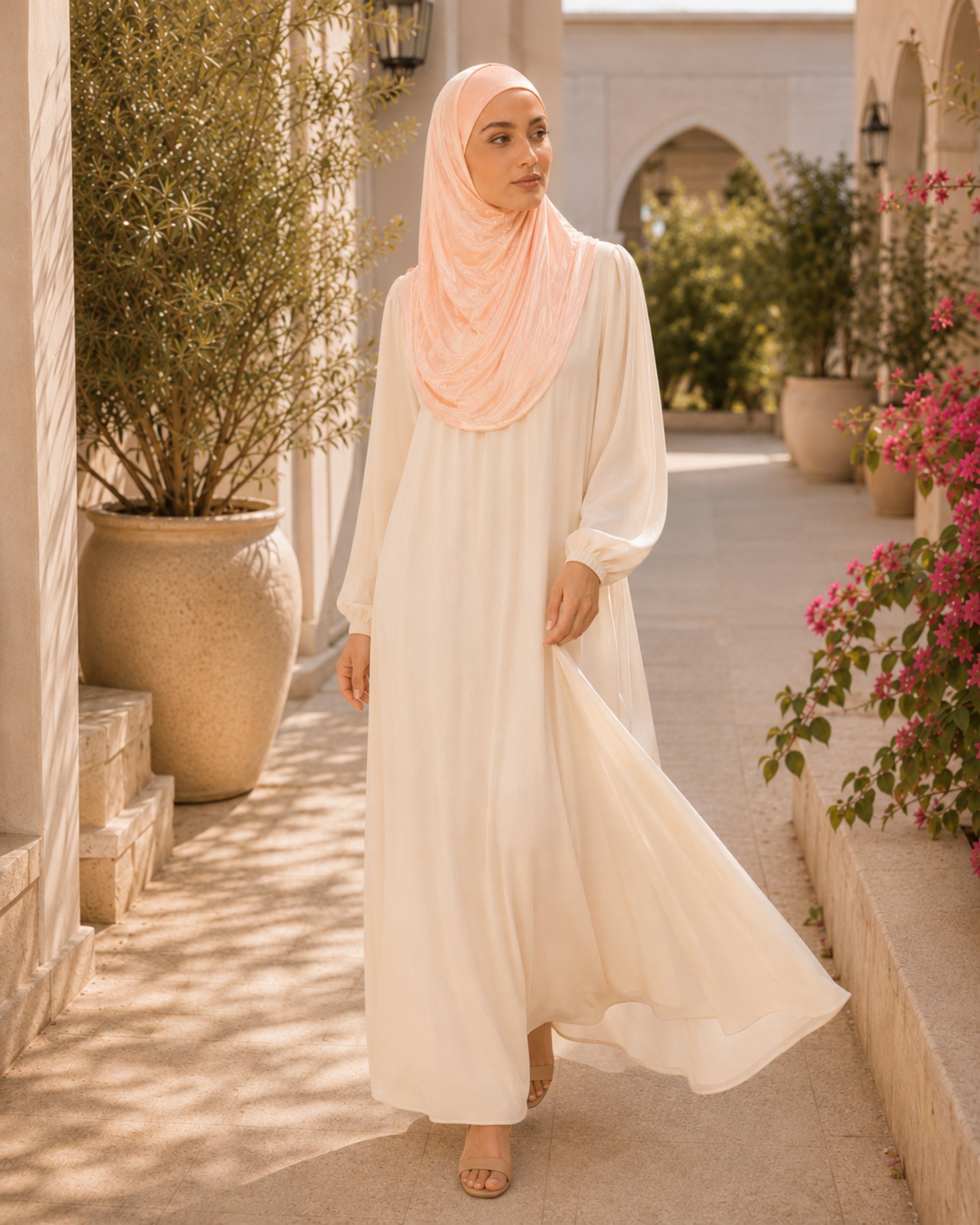 Woman in a long, light-colored dress and hijab standing in a sunlit outdoor setting with plants and flowers.

