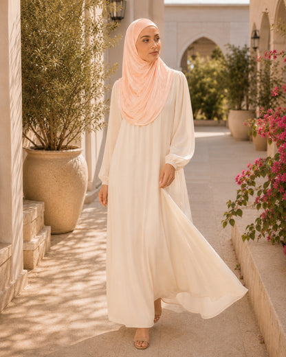 Woman in a long, light-colored dress and hijab standing in a sunlit outdoor setting with plants and flowers.

