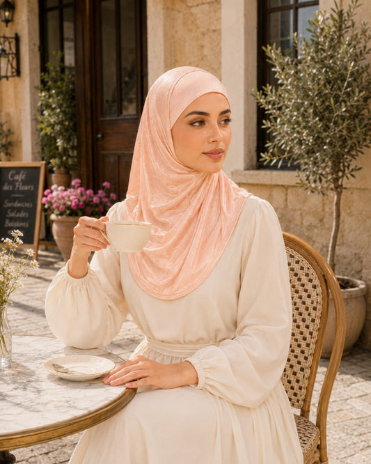 Woman in a hijab sitting at an outdoor cafe, holding a cup of coffee.

